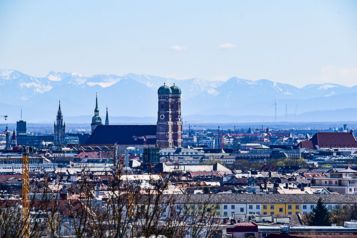 Blick auf die Altstadt von München mit der Frauenkirche mit ihren mächtigen Türmen mit Zwiebelkuppeln im Zentrum. Dahinter die Gipfel der Alpen