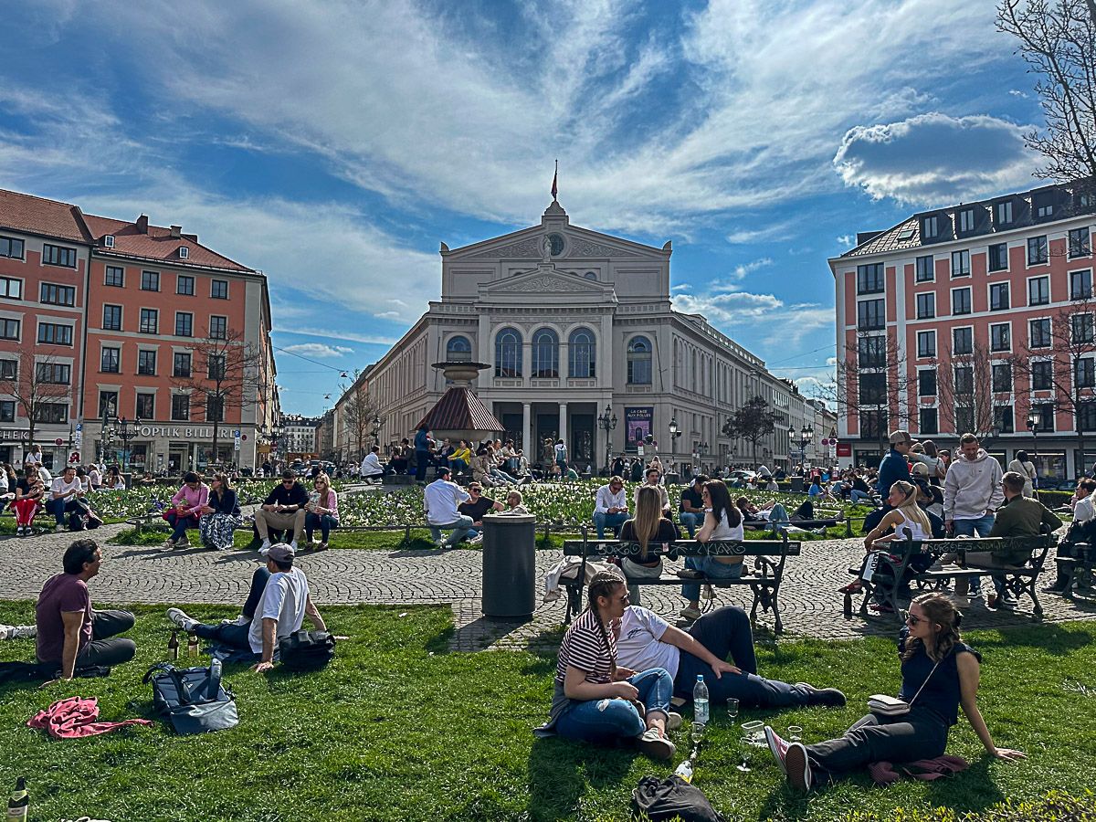 Menschen entspannen auf einer Rasenfläche und Bänken auf einem belebten Stadtplatz. Im Hintergrund ist ein prächtiges Gebäude unter blauem Himmel mit Wolken zu sehen. Die Szene ist lebhaft, und viele Menschen genießen die Natur.