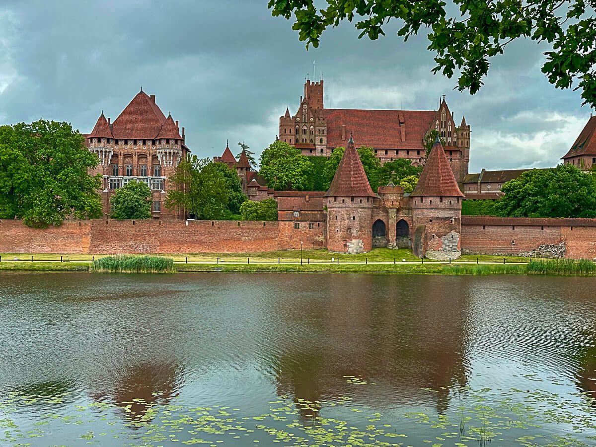 Malbork: eine Burg aus rotem Backstein erhebt sich am Flussufer. die Türme spiegeln sich im Wasser.