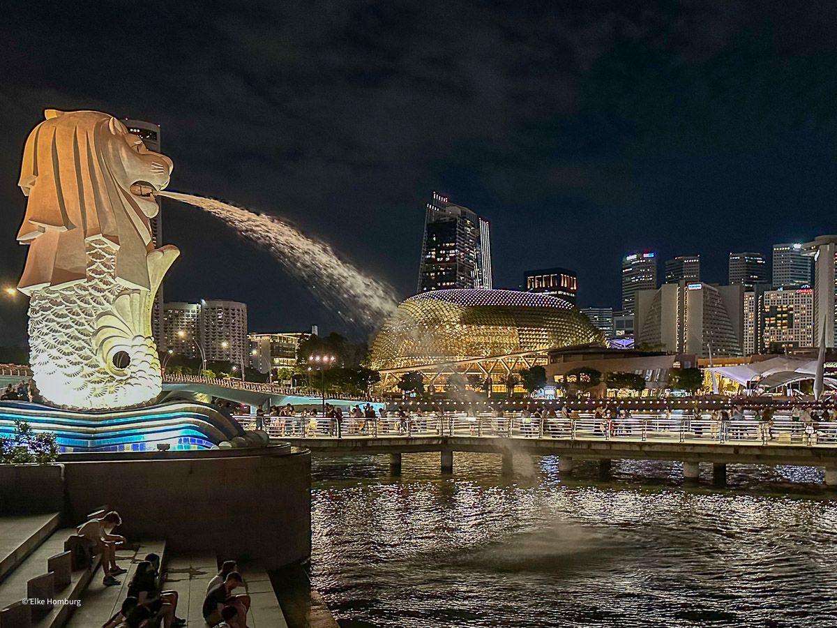 Merlion – das Maskottchen der Stadt – spukt abends auf die beleuchtete Stadt mit den Hochhäusern im Hintergrund