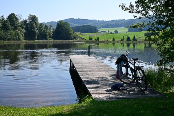 Radtouren im Pfaffenwinkel: ein Moorsee mit Steg, auf dem ein Fahrrad steht, vor oder Kulisse der Berge