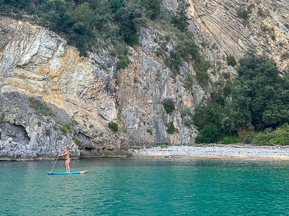 einsamer Strand, davor türkisfarbenes Wasser und eine Frau auf einem SUP
