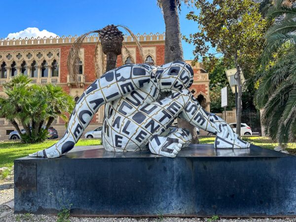 Highlights in Kalabrien: Moderne Skulptur einer kauernden Frau auf einem Podest. Im Hintergrund ein Palazzo.