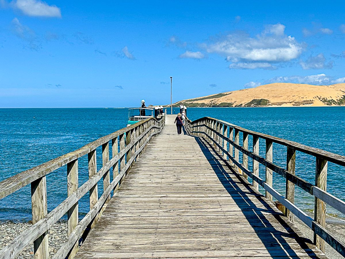 Hölzerne Brücke, die in eine Meeresbucht führt. Im Hintergrund eine Sanddüne.
