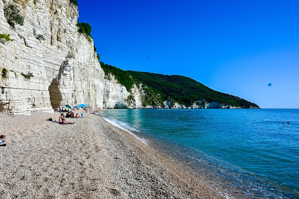 Kieselstrand vor weißem Fels mit ein paar Badegästen