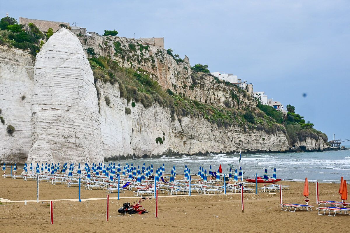 Ein weißer Kalkmonolith am Strand vor Liegestühlen, dahinter Vieste auf einem Fels