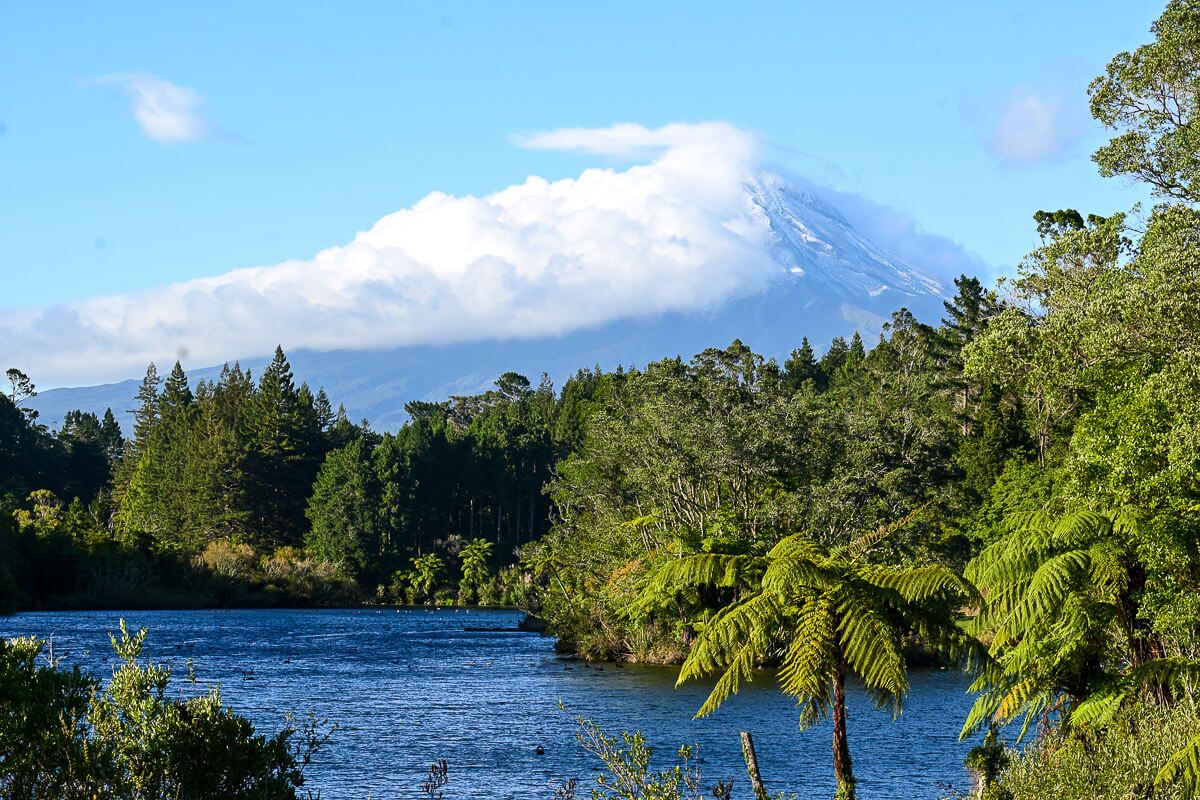 Neuseeland: Reisetipps für Mount Taranaki und New Plymouth 28 Blick über einen See im Wald auf den Vulkan Taranaki, der zum Teil in Wolken gehüllt ist und nur eine verschneite Flanke zeigt