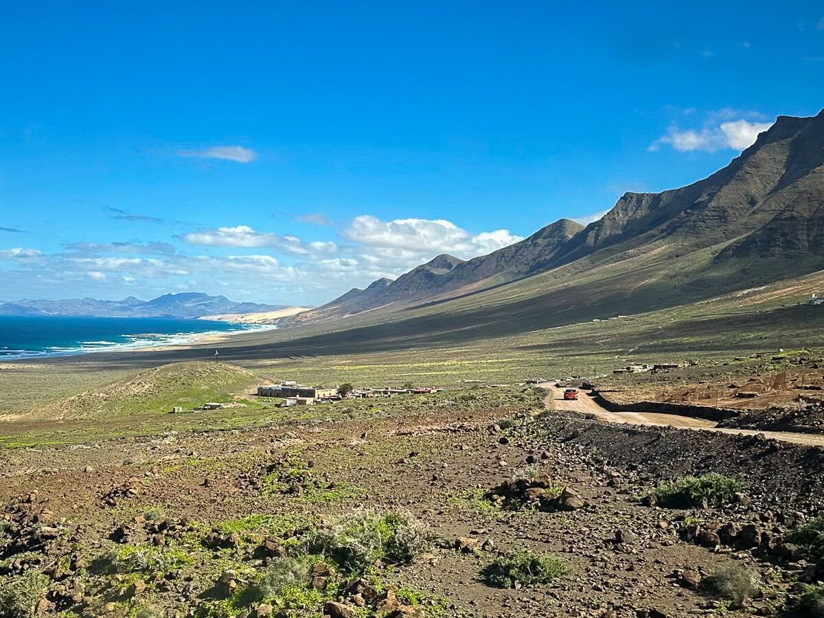 Blick auf eine wilde Küste. im Hintergrund ein paar verlassene Häuser und eine Piste mit einem Auto