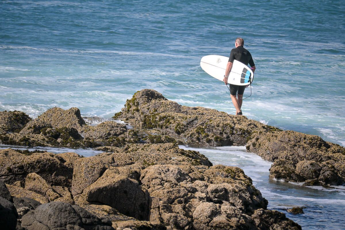 Surfer läuft mit seinem Brett über die Felsen in Manu Bay