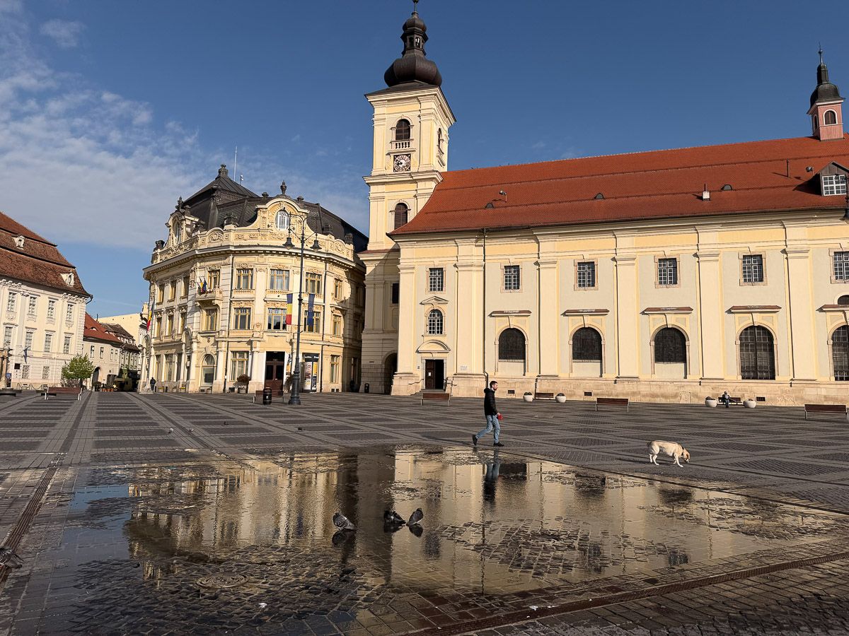 Großer Ring mit Katholischer Kirche, im Vordergrund spiegelt sich ein Haus in großer Pfütze