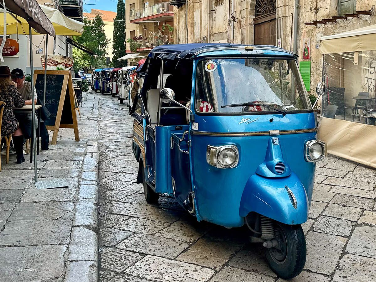 Viele Ape fahren hintereinander durch ein Stadtviertel